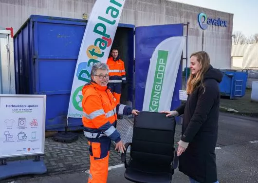 Een blauwe container van Rataplan kringloop, waar een oudere mannelijke medewerker een zwarte bureau stoel geeft aan een jonge vrouw in een zwarte jas.